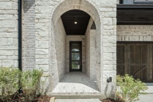 A house entryway with a white brick arch, glass front door, and a small garden with shrubs on either side of the walkway—perfect for those seeking beautiful homes for sale in McKinney, Texas.
