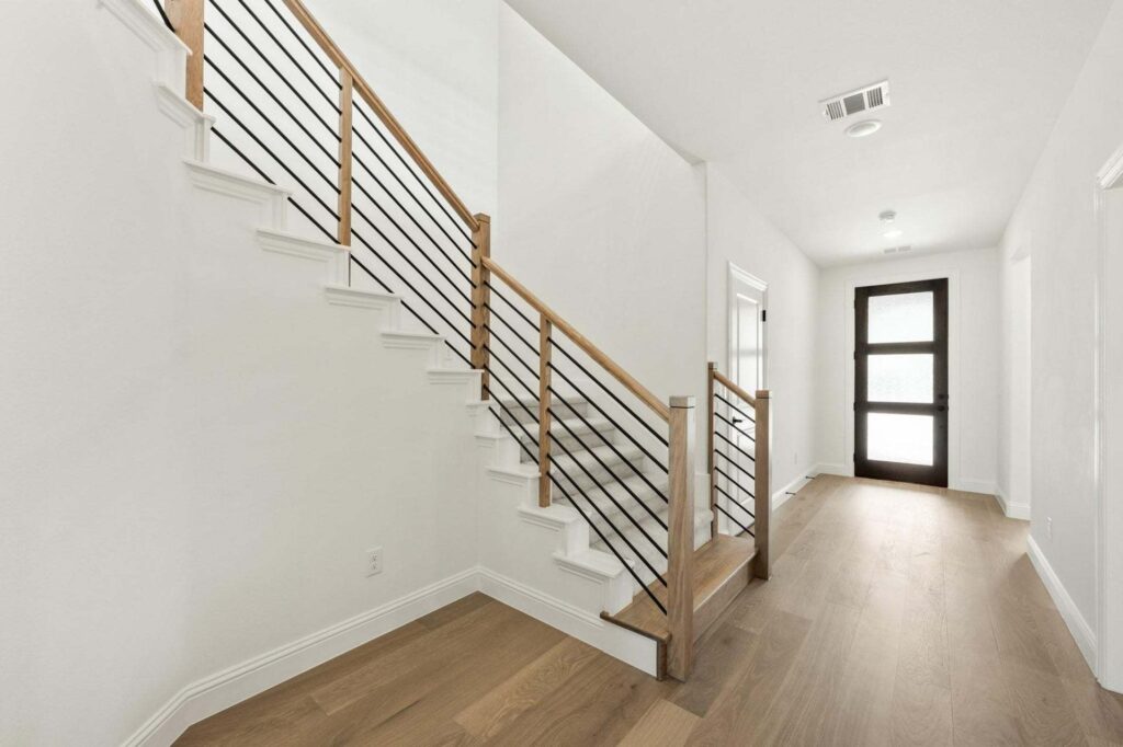 A modern hallway with light wood floors, white walls, a staircase with metal railings and wood handrail, and a glass front door—an inviting entry often found in homes for sale in McKinney, Texas.