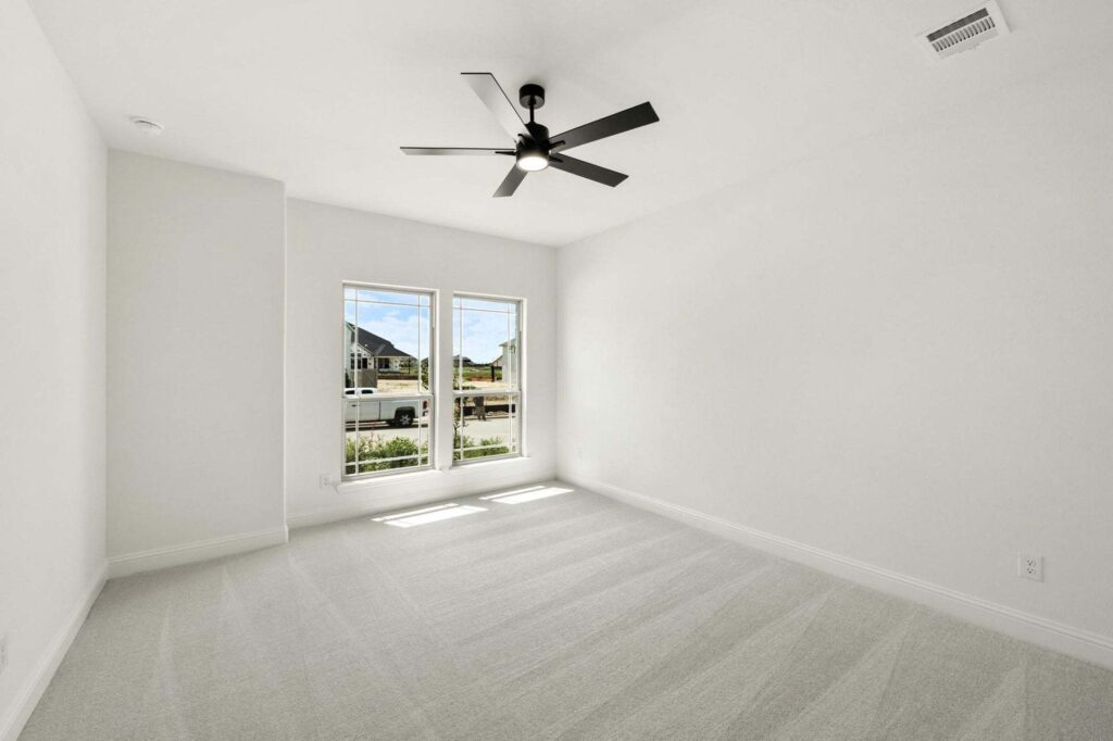 Empty, carpeted bedroom with white walls in a Painted Tree McKinney home, featuring two large windows that fill the space with natural light and a sleek black ceiling fan centered on the ceiling.