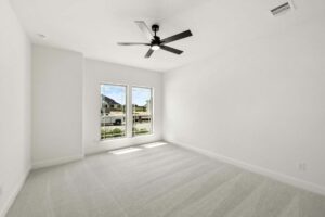 Empty, carpeted bedroom with white walls in a Painted Tree McKinney home, featuring two large windows that fill the space with natural light and a sleek black ceiling fan centered on the ceiling.