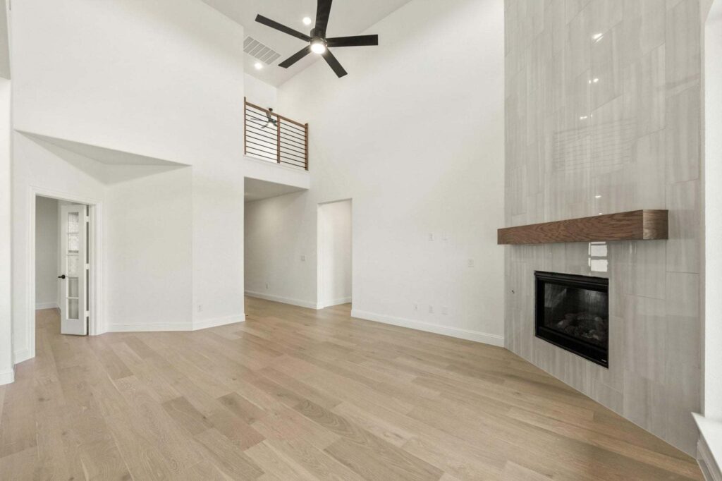 Spacious modern living room in one of the stunning Painted Tree TX homes for sale, featuring light wood floors, high ceilings, a wood mantel fireplace, ceiling fan, and a loft area with railing.