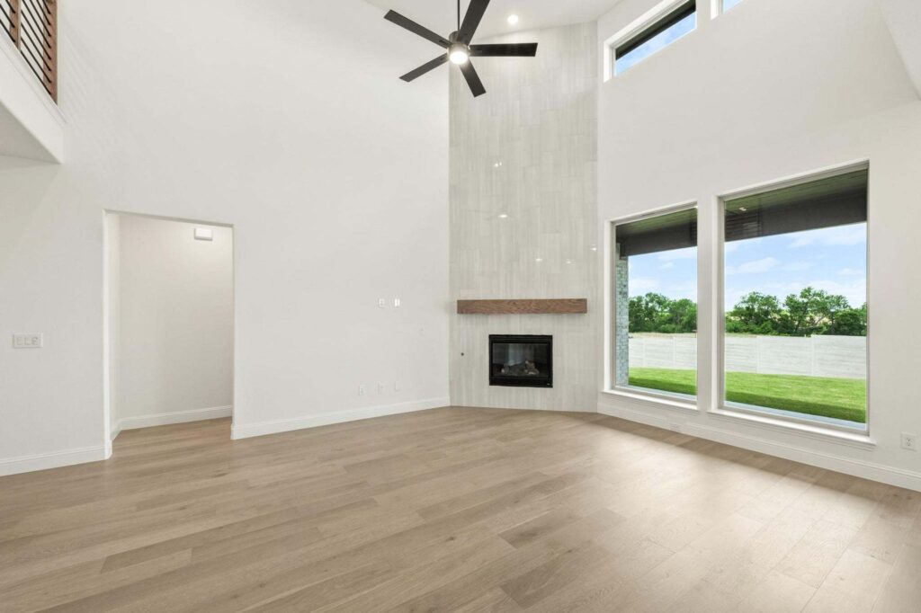 Modern living room in Painted Tree McKinney with light wood floors, tall white walls, large windows, high ceiling, a ceiling fan, and a fireplace featuring a wood mantel beneath vertical tiles. Explore homes for sale in McKinney Texas today.