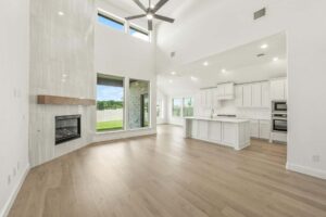 Modern open-concept living area with light wood flooring, tall ceilings, large windows, a fireplace, and a white kitchen with an island—perfect for anyone searching for stylish homes for sale in McKinney Texas.