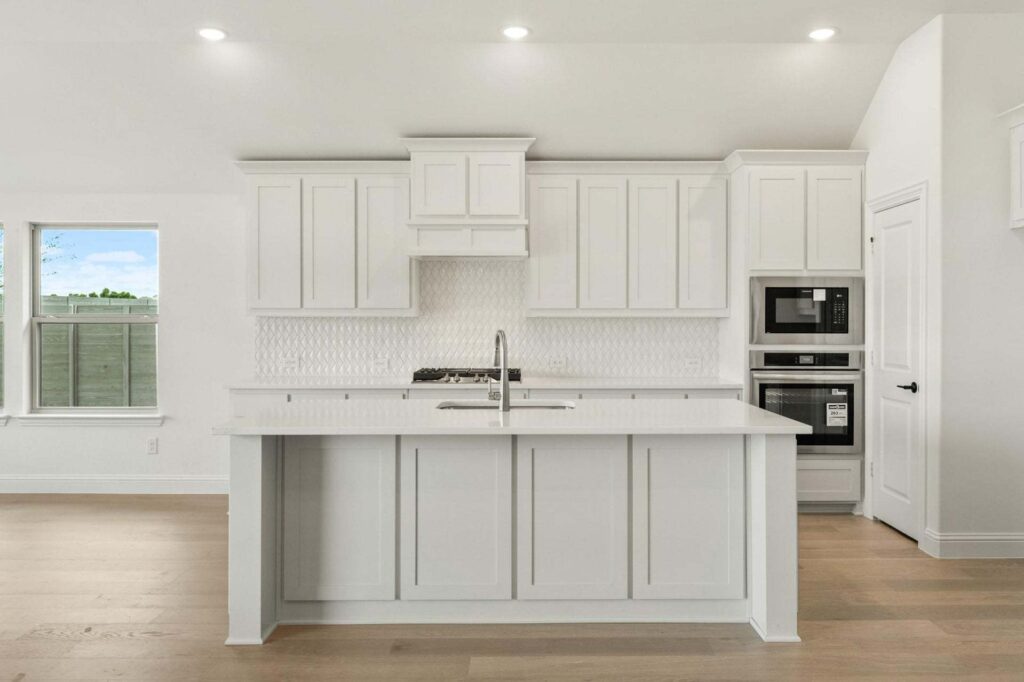Modern kitchen in Painted Tree McKinney features white cabinets, a center island with sink, built-in microwave and oven, light wood floors, and a patterned tile backsplash—perfect for those seeking homes for sale in McKinney, Texas.