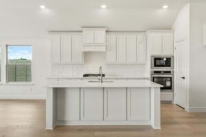 Modern kitchen in Painted Tree McKinney features white cabinets, a center island with sink, built-in microwave and oven, light wood floors, and a patterned tile backsplash—perfect for those seeking homes for sale in McKinney, Texas.