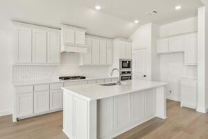 Modern kitchen in Painted Tree McKinney featuring white cabinets, white countertops, built-in oven, gas stove, tile backsplash, and a central island with sink. Enjoy light wood flooring and recessed lighting throughout.