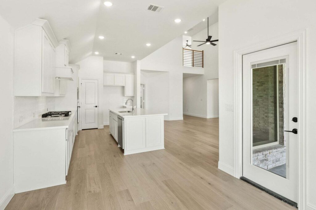 Modern kitchen and open living area with white cabinets, island, light wood flooring, high ceiling, and glass door leading outside—discover this inviting space among homes for sale in McKinney Texas.