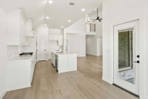 Modern kitchen and open living area with white cabinets, island, light wood flooring, high ceiling, and glass door leading outside—discover this inviting space among homes for sale in McKinney Texas.