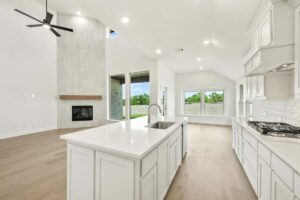 Modern open-plan kitchen and living area with white cabinetry, island, built-in fireplace, large windows, wood floors, and high ceilings—perfect for those seeking Painted Tree McKinney homes for sale.