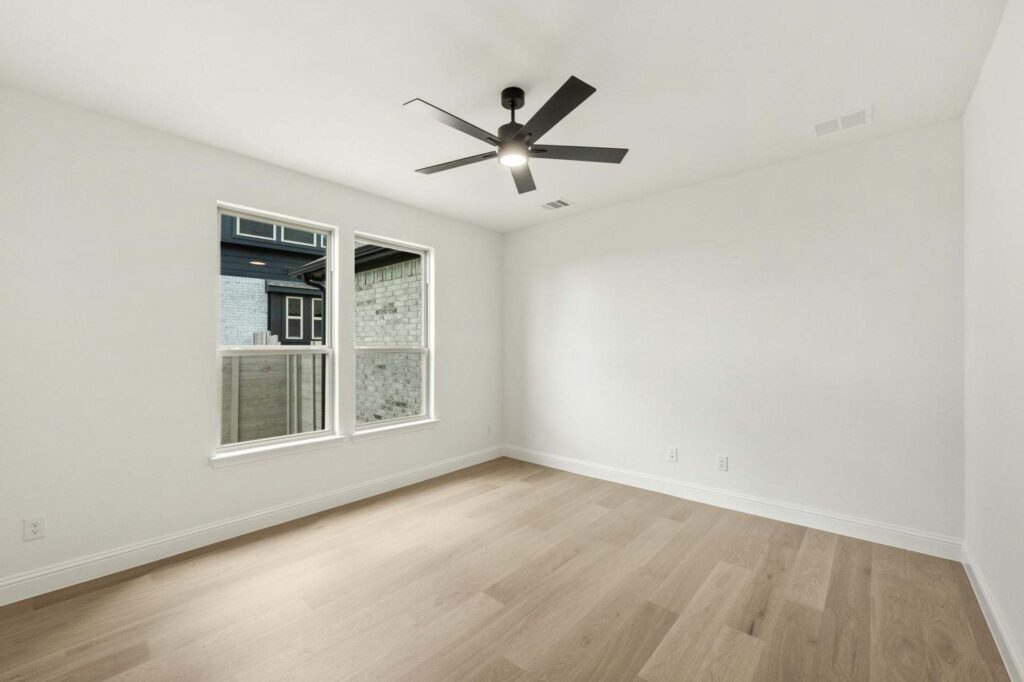 Empty room with light wood flooring, white walls, two windows, ceiling vent, and a sleek black modern ceiling fan—perfect for those seeking Painted Tree McKinney homes for sale or elegant living in Painted Tree TX.