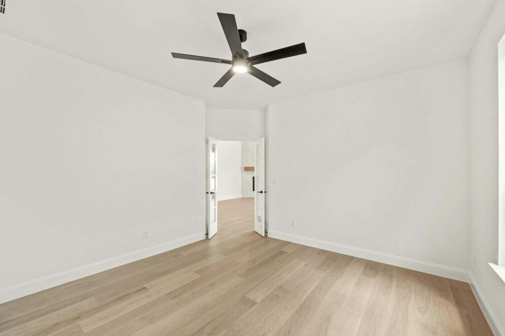 Bright empty room with light wood flooring, white walls, a black ceiling fan, double doors, and a large window on the right—perfect for your next home at Painted Tree McKinney.