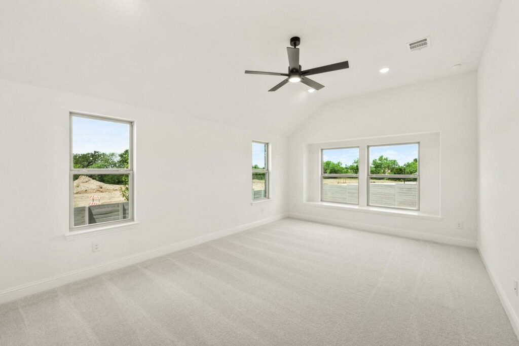 Empty, bright room with light-colored carpet, white walls, three large windows, and a black ceiling fan. Enjoy views of green trees and a construction area—ideal for those exploring painted tree homes for sale in McKinney, Texas.