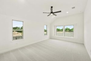 Empty, bright room with light-colored carpet, white walls, three large windows, and a black ceiling fan. Enjoy views of green trees and a construction area—ideal for those exploring painted tree homes for sale in McKinney, Texas.