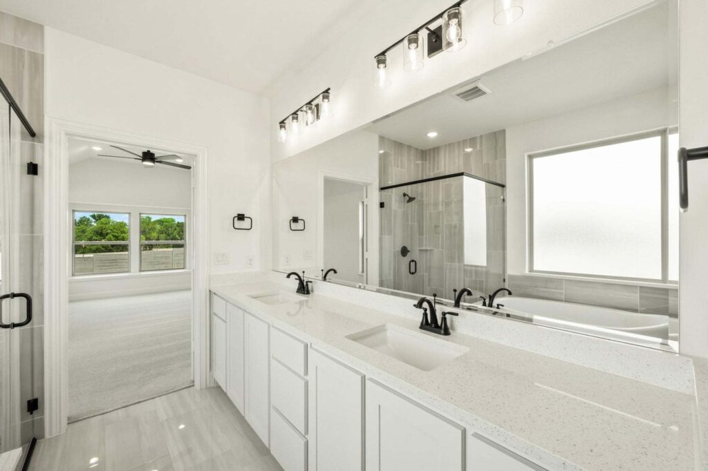 Modern bathroom with double sinks, white cabinets, large mirror, black fixtures, glass-enclosed shower, and a frosted window. Bedroom visible through open door in this stylish Painted Tree McKinney home.