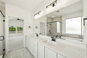 Modern bathroom with double sinks, white cabinets, large mirror, black fixtures, glass-enclosed shower, and a frosted window. Bedroom visible through open door in this stylish Painted Tree McKinney home.