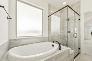 Bathroom in a Painted Tree McKinney home featuring a large oval bathtub, glass-enclosed shower, frosted window, and elegant gray tile. Black fixtures accent the tub and shower—discover stylish homes for sale in McKinney, Texas.