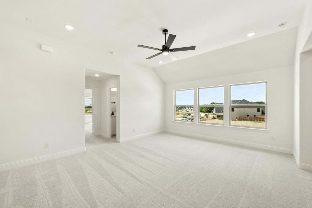Bright, empty room with light carpet, large windows showing a neighborhood view in Painted Tree McKinney, ceiling fan, recessed lighting; doorway leads to another area.