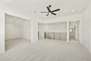 A spacious, carpeted upstairs loft area with white walls, a modern ceiling fan, recessed lighting, and a metal railing overlooking stairs—perfect for relaxing in one of the stunning Painted Tree homes for sale in McKinney, Texas.