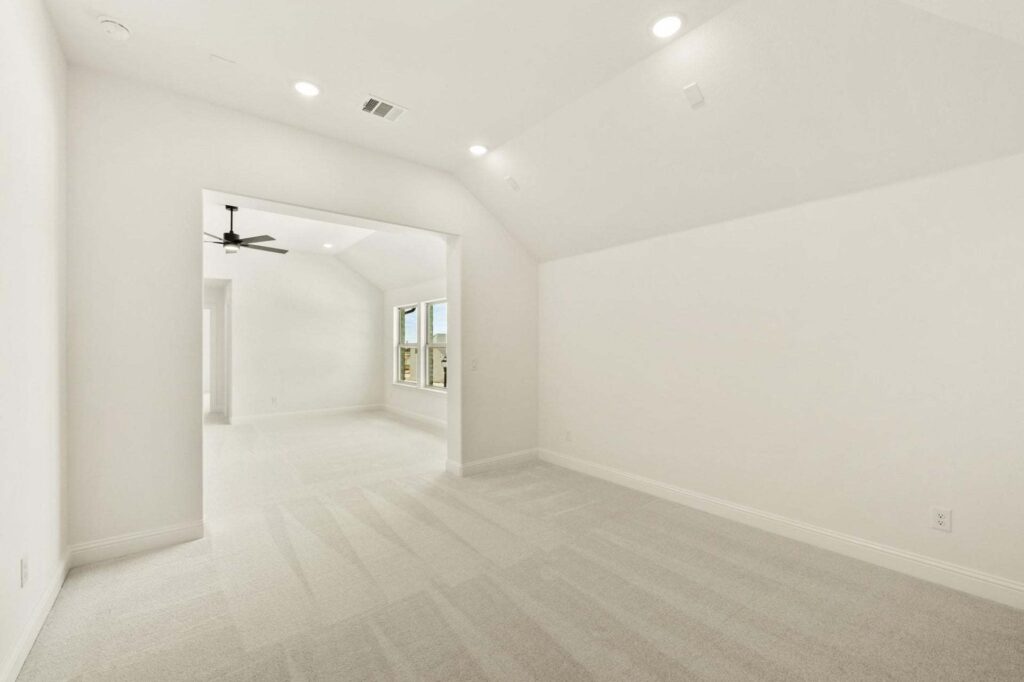A spacious empty room with light-colored carpet, white walls, recessed lighting, and an open doorway leading to another area with a ceiling fan and windows—perfect for those seeking Painted Tree McKinney homes for sale.
