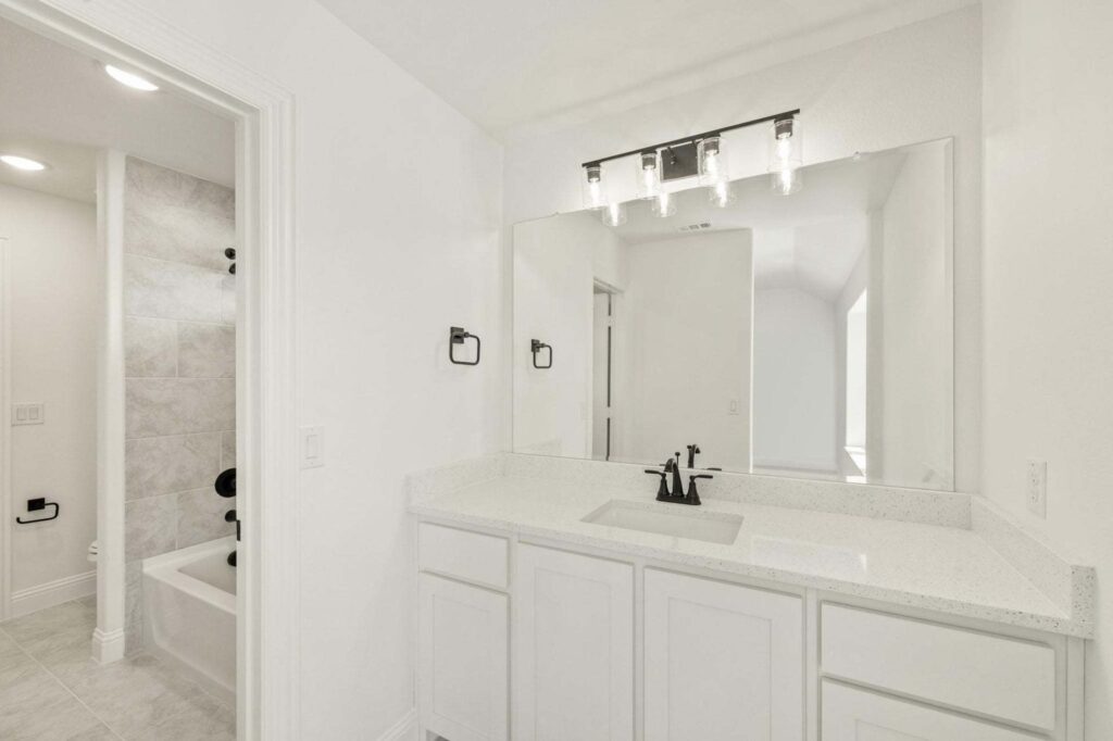 Modern bathroom with white cabinets, quartz countertop, large mirror, black fixtures, and a view of a bathtub with tiled walls in the adjacent room—perfect for those seeking stylish comfort in Painted Tree McKinney.