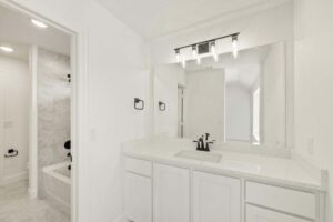 Modern bathroom with white cabinets, quartz countertop, large mirror, black fixtures, and a view of a bathtub with tiled walls in the adjacent room—perfect for those seeking stylish comfort in Painted Tree McKinney.