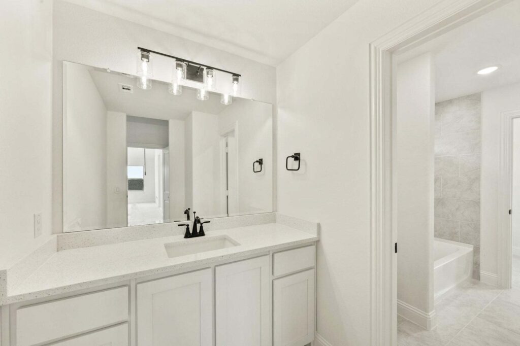 Bathroom with white cabinets, quartz countertop, single sink, black faucet and hardware, large wall mirror, and a view into a tiled bathtub area in one of the beautiful Painted Tree TX homes for sale.