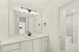 Bathroom with white cabinets, quartz countertop, single sink, black faucet and hardware, large wall mirror, and a view into a tiled bathtub area in one of the beautiful Painted Tree TX homes for sale.