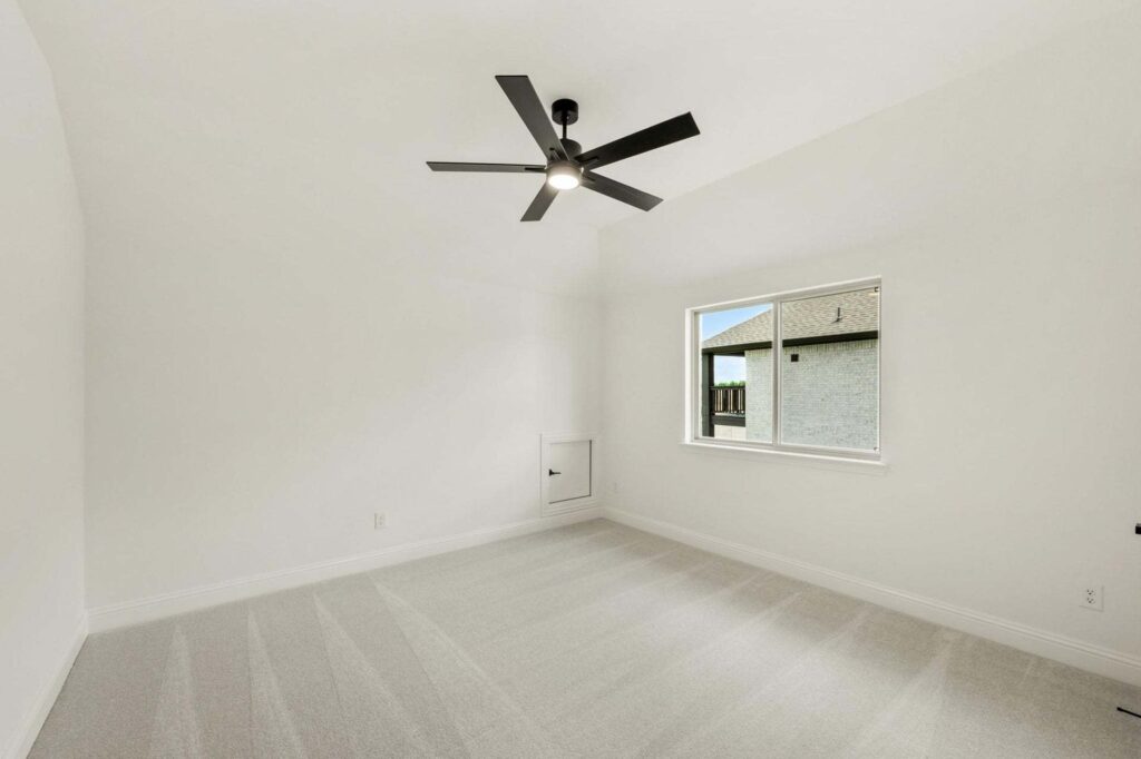 Empty room with white walls, light-colored carpet, black ceiling fan with light, a window, and a small wall hatch near the floor—perfect for those searching painted tree homes for sale in this desirable community.