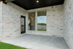 A covered patio with a concrete floor, two large windows, a black-framed glass door, and light-colored brick exterior walls—perfect for those seeking Painted Tree homes for sale in McKinney Texas.
