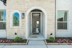 Arched front entrance of a light brick house with two windows, flower beds, and a glass door leading to the interior hallway—one of the inviting homes for sale in McKinney, Texas.