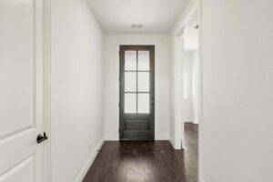 A hallway with dark wood flooring, white walls, and a black framed glass door at the end; doorways to other rooms are visible on the right in this stunning Painted Tree TX home.