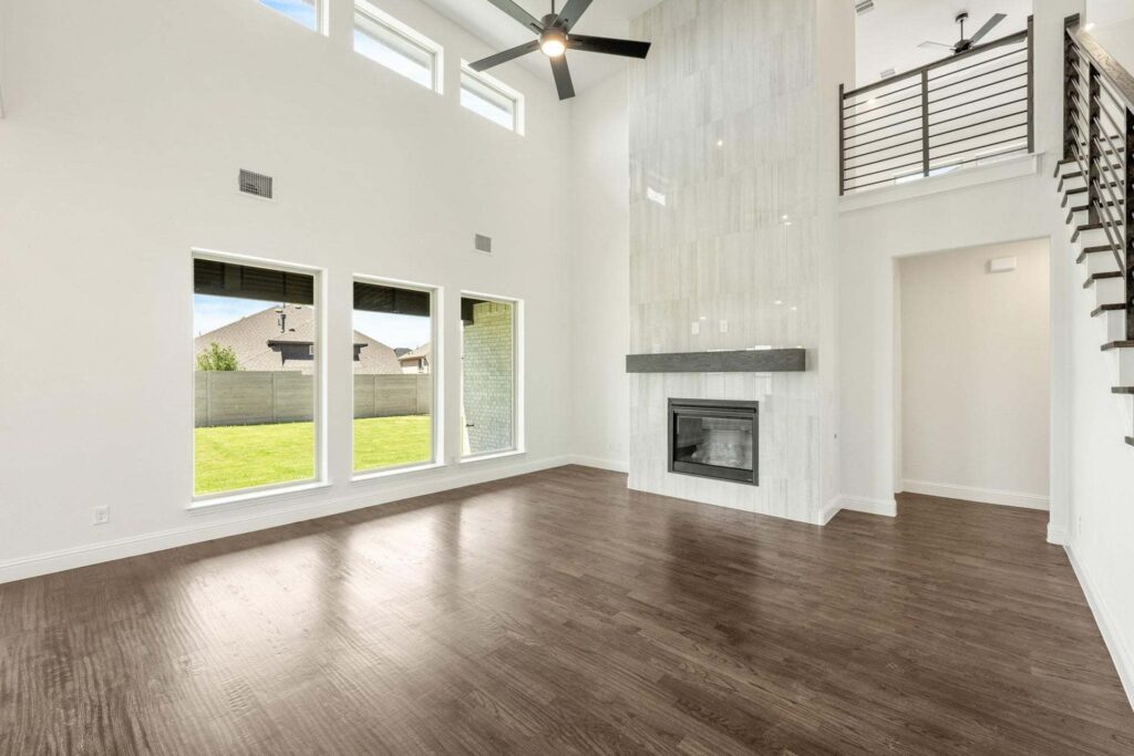 Spacious living room with high ceiling, large windows, dark wood floors, modern fireplace, ceiling fan, and a metal-railed staircase—discover homes for sale in McKinney Texas like this in the beautiful Painted Tree community.