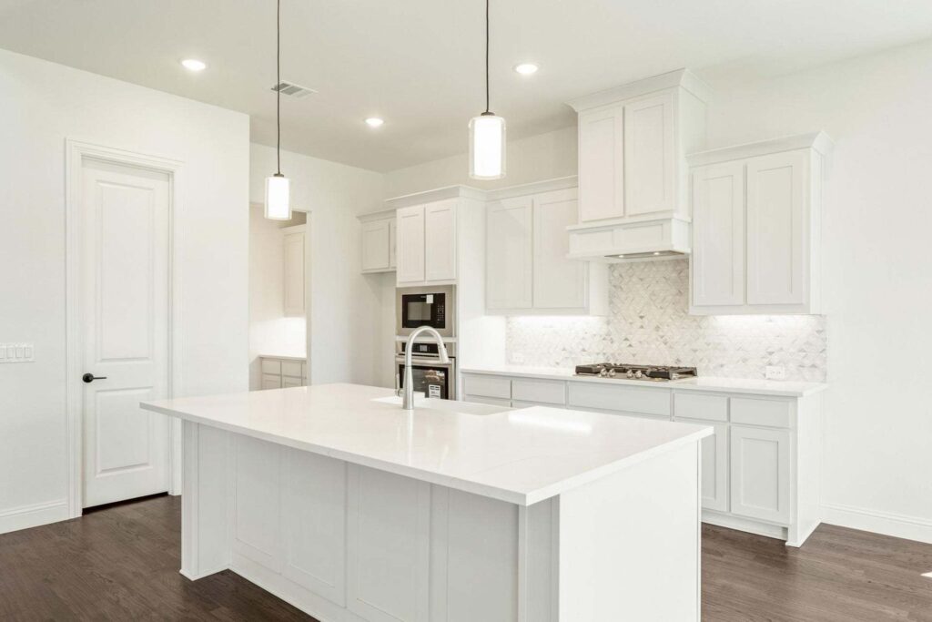 Modern kitchen with white cabinets, a large island with sink, pendant lights, built-in oven, stovetop, and marble backsplash on dark hardwood floors—an elegant feature found in many Painted Tree McKinney homes for sale.