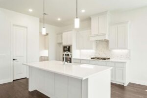 Modern kitchen with white cabinets, a large island with sink, pendant lights, built-in oven, stovetop, and marble backsplash on dark hardwood floors—an elegant feature found in many Painted Tree McKinney homes for sale.