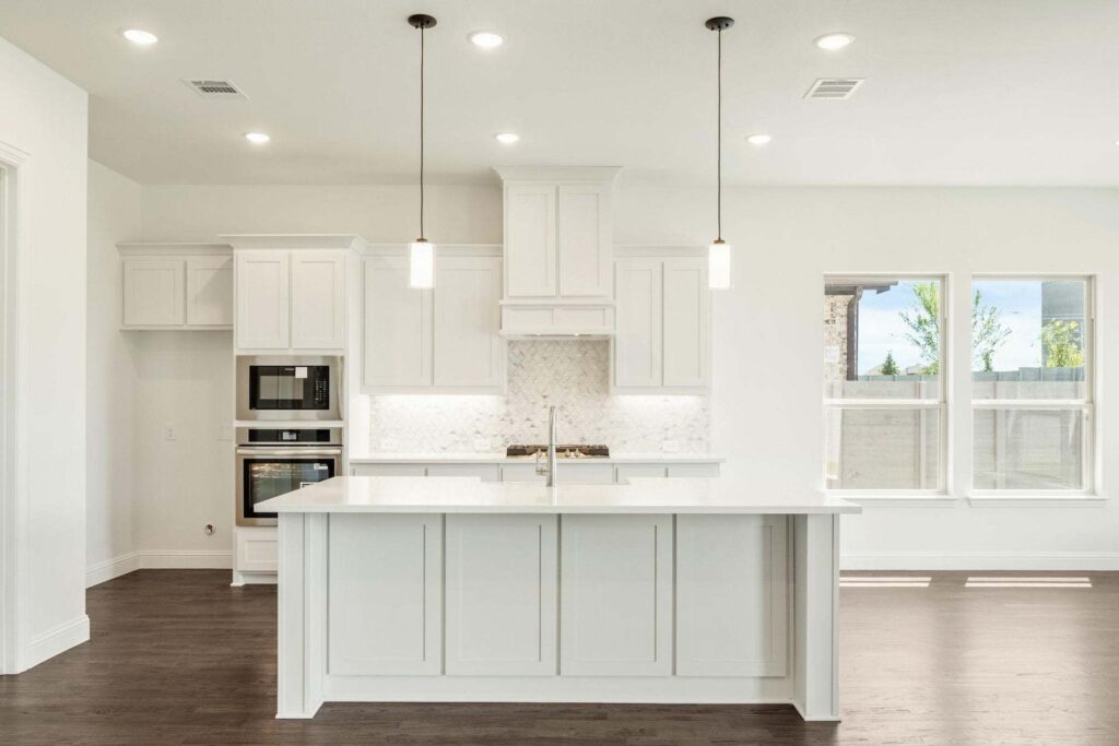 Modern kitchen in Painted Tree McKinney with white cabinets, a spacious island with sink, built-in oven, pendant lights, and dark wood floors. Ample natural light streams through large windows on the right.