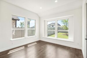 Bright, empty room with white walls, large windows, and dark wood floors, overlooking a fenced backyard with grass and a tree in Painted Tree McKinney.