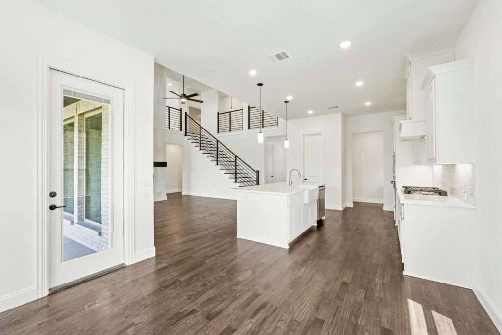 Modern open-concept kitchen and living area with white cabinets, island, wood flooring, staircase with metal railing, and glass door leading outside—perfect for those seeking homes for sale in McKinney Texas.