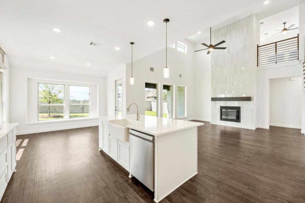 Modern open-concept living space in Painted Tree McKinney features a white kitchen island, stainless steel sink, pendant lights, fireplace, large windows, and dark wood flooring. Discover stylish living with Painted Tree homes for sale.