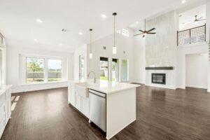 Modern open-concept living space in Painted Tree McKinney features a white kitchen island, stainless steel sink, pendant lights, fireplace, large windows, and dark wood flooring. Discover stylish living with Painted Tree homes for sale.