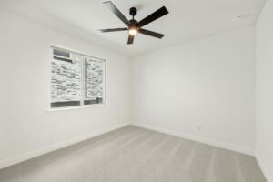 Empty, unfurnished room with white walls, light gray carpet, a large window, and a sleek black ceiling fan—discover this inviting space among Painted Tree homes for sale in McKinney, Texas.