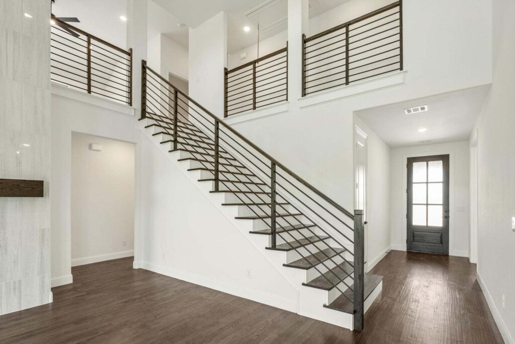 Modern interior with dark wood flooring, open staircase with metal railing, high ceilings, and a hallway leading to a glass-paneled front door—perfect for those seeking Painted Tree McKinney style and elegance.