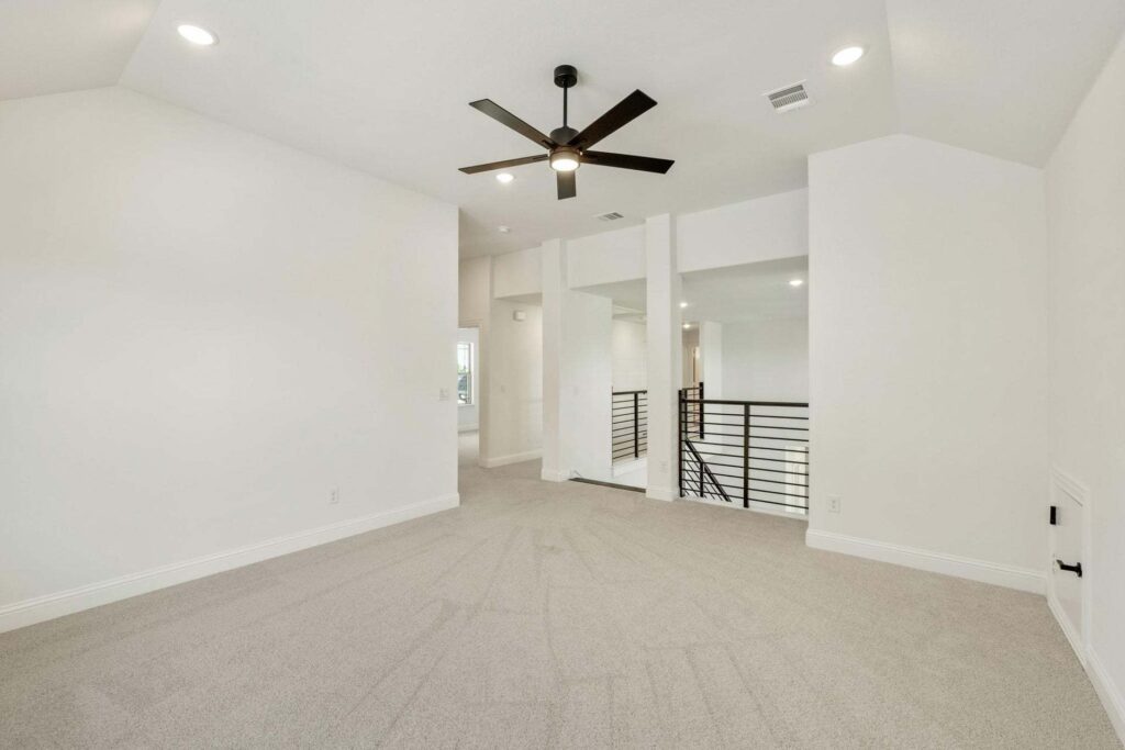 Spacious, empty room with light beige carpet, white walls, black ceiling fan, recessed lighting, and metal railing overlooking a lower level—perfect for those seeking Painted Tree McKinney homes for sale.