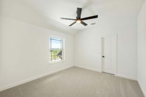 Empty room with white walls, beige carpet, ceiling fan with light, single window showing an outdoor view, and closed white door—discover tranquil spaces like this in Painted Tree McKinney when searching for homes for sale in McKinney Texas.