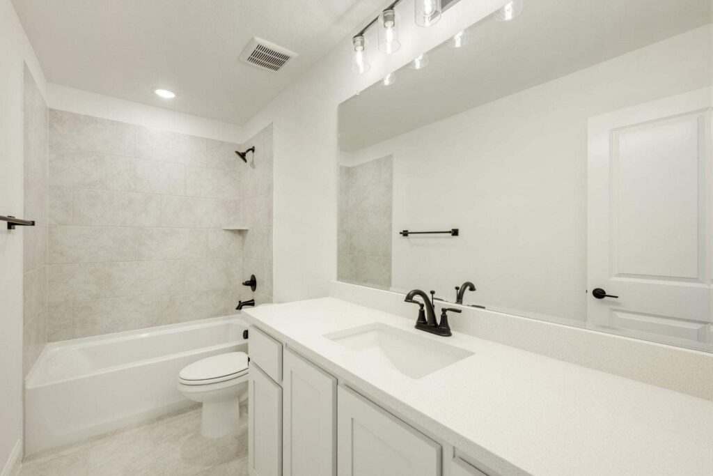 Modern white bathroom in a Painted Tree McKinney home, featuring a bathtub, toilet, large mirror, sink, black fixtures, and tiled shower walls. The room is well-lit with ceiling and vanity lights.
