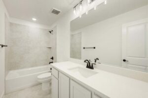 Modern white bathroom in a Painted Tree McKinney home, featuring a bathtub, toilet, large mirror, sink, black fixtures, and tiled shower walls. The room is well-lit with ceiling and vanity lights.