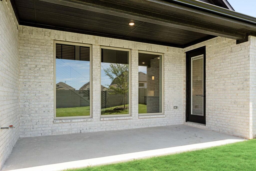 A covered patio with a concrete floor, white brick walls, three windows, and a glass door overlooks a grassy backyard—perfect for relaxing in one of the beautiful Painted Tree homes for sale.