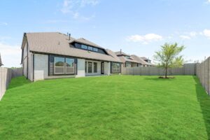 Back view of a modern two-story house in Painted Tree McKinney with large windows, covered patio, fenced yard, and a single tree on a well-maintained lawn under a partly cloudy sky. Explore Painted Tree homes for sale today!.