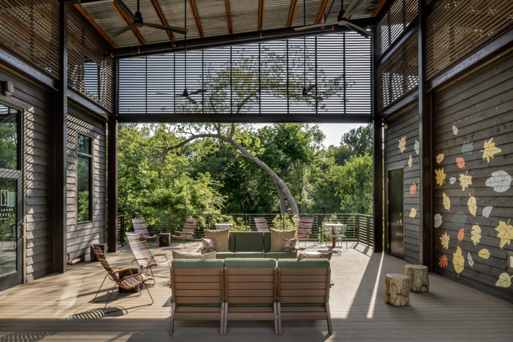 A covered outdoor deck at The Outpost offers wooden seating, metal chairs, and a view of trees. Painted Tree-inspired leaf designs adorn the wall; large windows and a slatted roof fill this amenity with natural light.