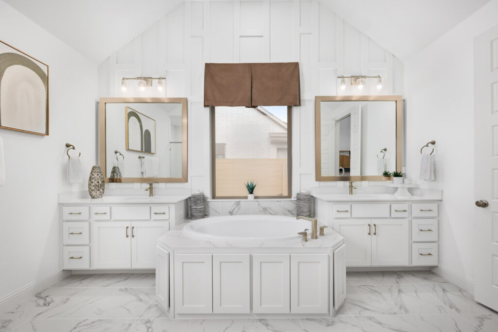 Modern bathroom in a new home at Painted Tree, McKinney, featuring a central bathtub, dual vanities, large mirrors, marble floors, and a window with a brown roman shade above the tub.