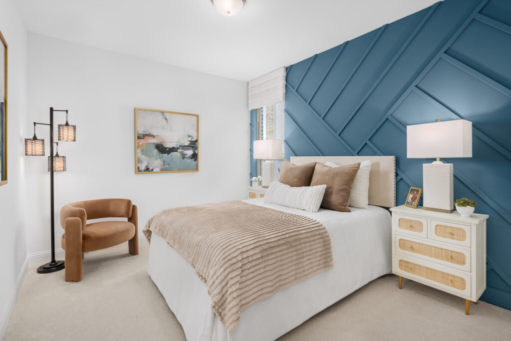 A modern bedroom by Coventry Homes in McKinney, TX features a blue accent wall, beige bedding, two nightstands with lamps, a brown chair, and abstract artwork above the chair.